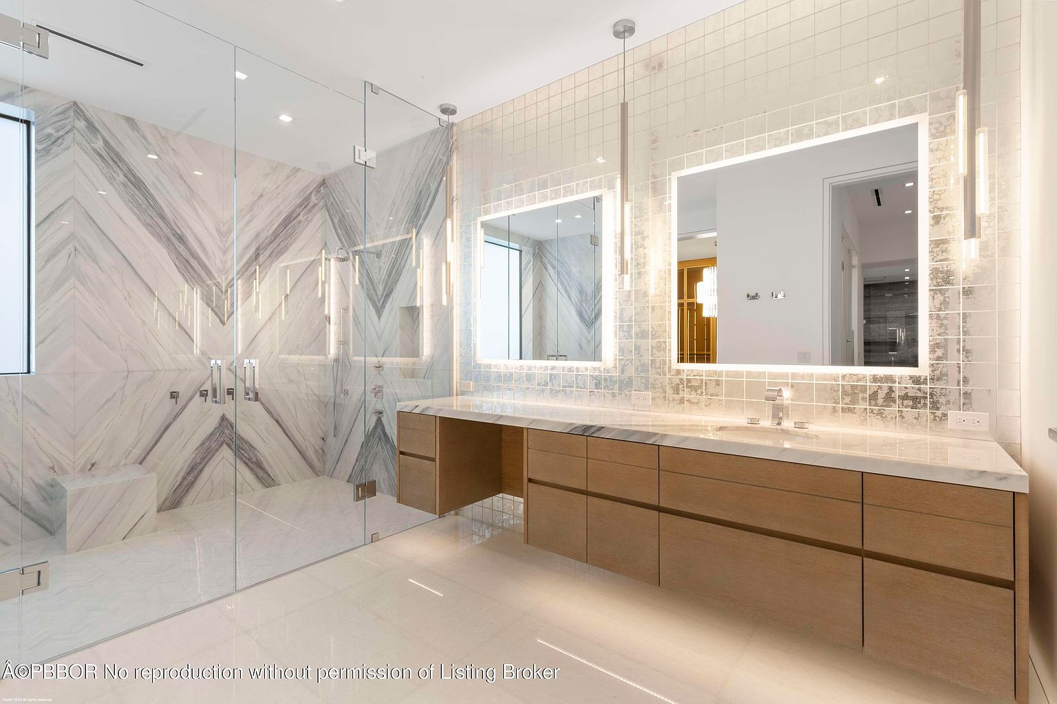 This is a luxurious primary bathroom featuring a glass-enclosed shower with marble chevron patterned walls and a built-in bench. The vanity area includes a long countertop with double sinks, light wood cabinetry, and illuminated mirrors. The overall design is modern and elegant, creating a spa-like atmosphere.