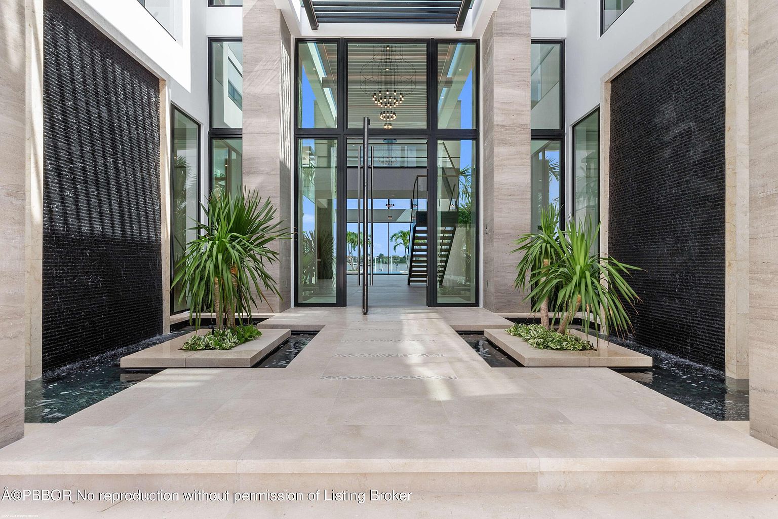 This is a grand entryway to a modern home, featuring a striking water feature on either side of the walkway leading to large glass doors. The architecture is characterized by clean lines, travertine stone accents, and lush greenery, creating an inviting and luxurious first impression. The open doors reveal a glimpse of the interior staircase and chandelier.