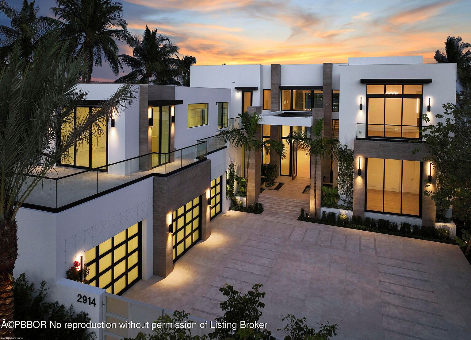 This is a front exterior view of a modern, two-story home at dusk. The house features a white facade with dark-framed windows and doors, accented by stone columns and a flat roof. A spacious driveway leads to the garage, and palm trees add a tropical touch to the landscaping. The overall impression is one of luxury and contemporary design.