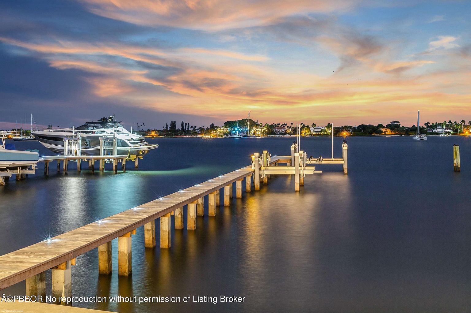 This stunning rear view showcases a luxurious waterfront property at dusk. A private dock extends into the calm water, featuring a modern yacht and additional boat lifts. The sky is painted with vibrant hues of orange and blue, reflecting on the water's surface, creating a serene and upscale ambiance.
