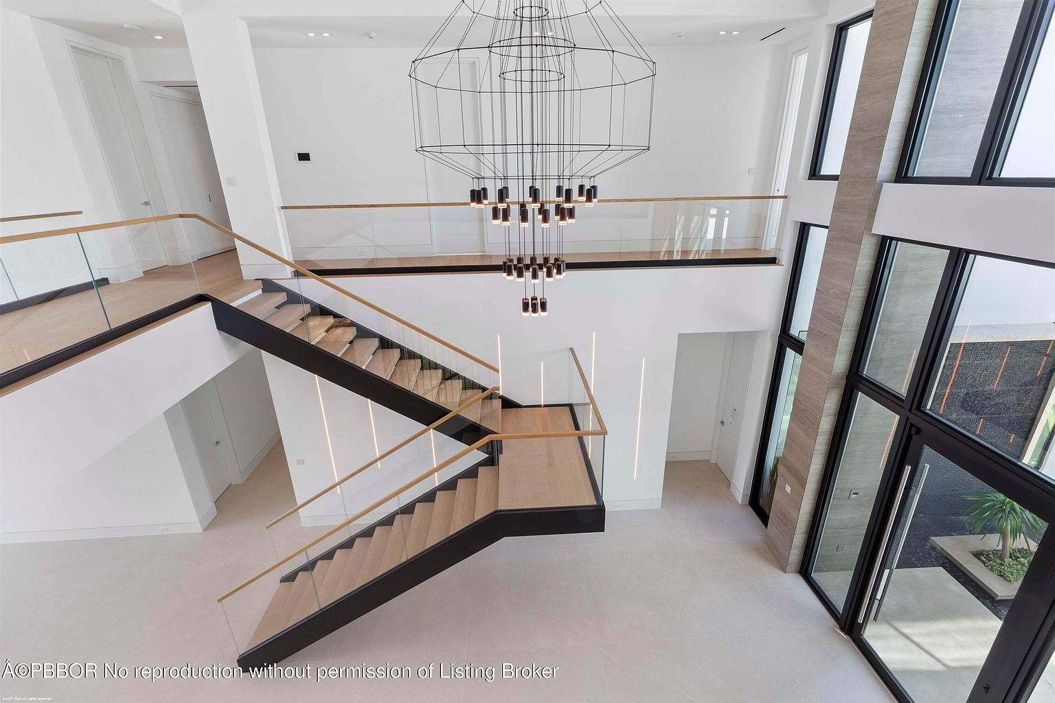 This is an interior shot of a modern home's grand foyer, showcasing a striking staircase with wooden steps, black accents, and glass railings. A large, geometric chandelier hangs above, adding a dramatic focal point. The space is bright and airy, with clean white walls and large windows that let in plenty of natural light, creating a luxurious and inviting atmosphere.