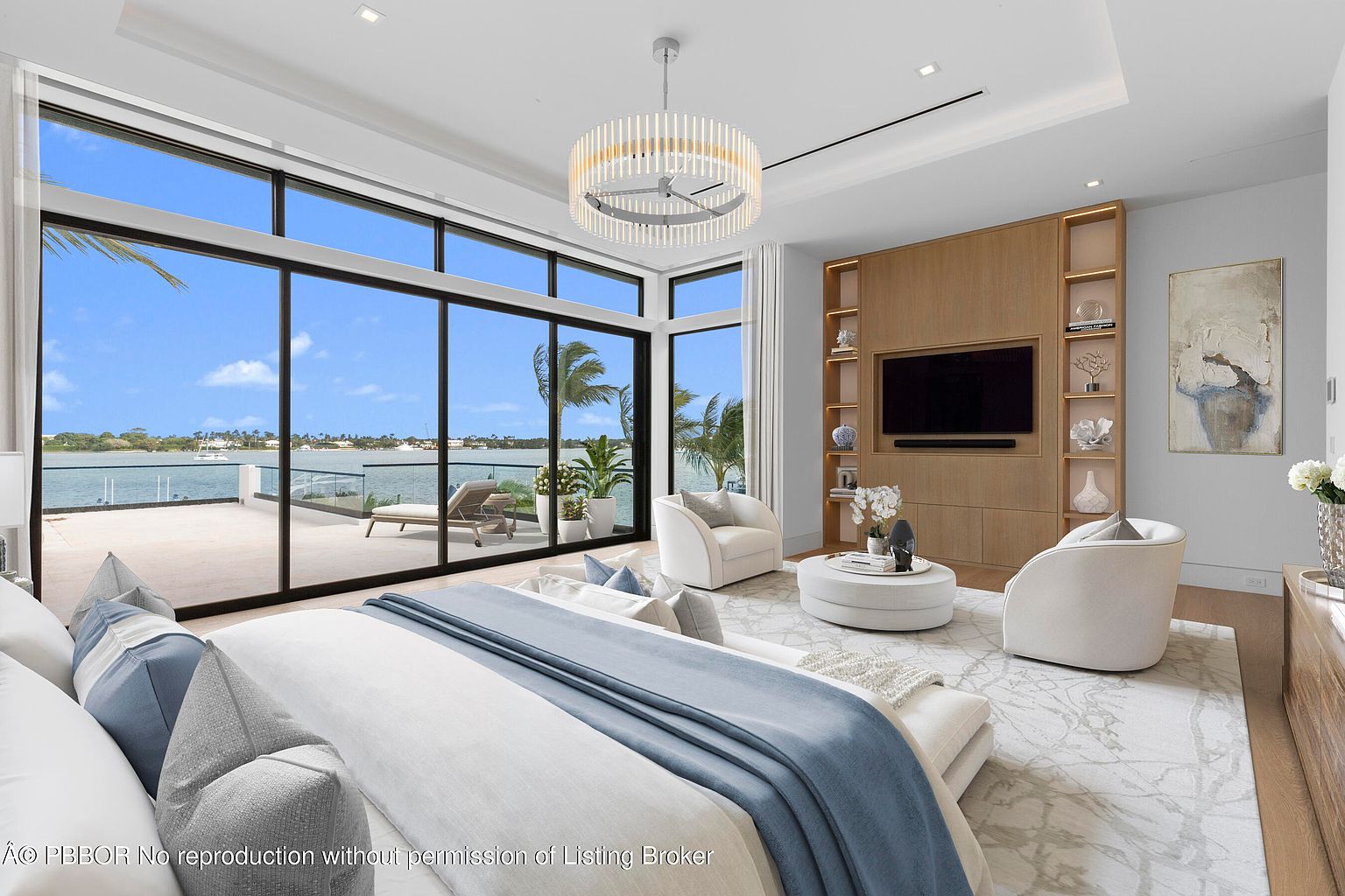This is a luxurious primary bedroom featuring a large bed with blue accents, floor-to-ceiling windows offering water views, and a seating area with two armchairs and a round coffee table. A built-in entertainment center with shelving and a large television adds to the room's appeal. The overall style is modern and elegant, creating a serene and inviting atmosphere.