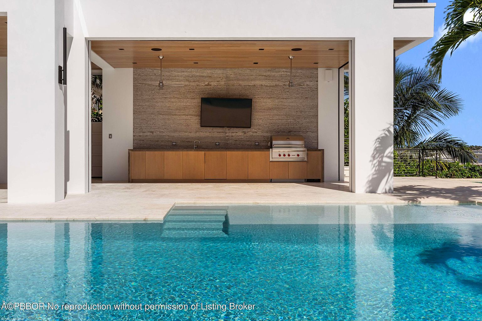 This image showcases a luxurious outdoor pool area with a modern outdoor kitchen. The pool's clear blue water contrasts with the light-colored stone patio and the sleek, contemporary design of the kitchen area, which includes a built-in grill and cabinetry. The scene evokes a sense of relaxation and upscale outdoor living.