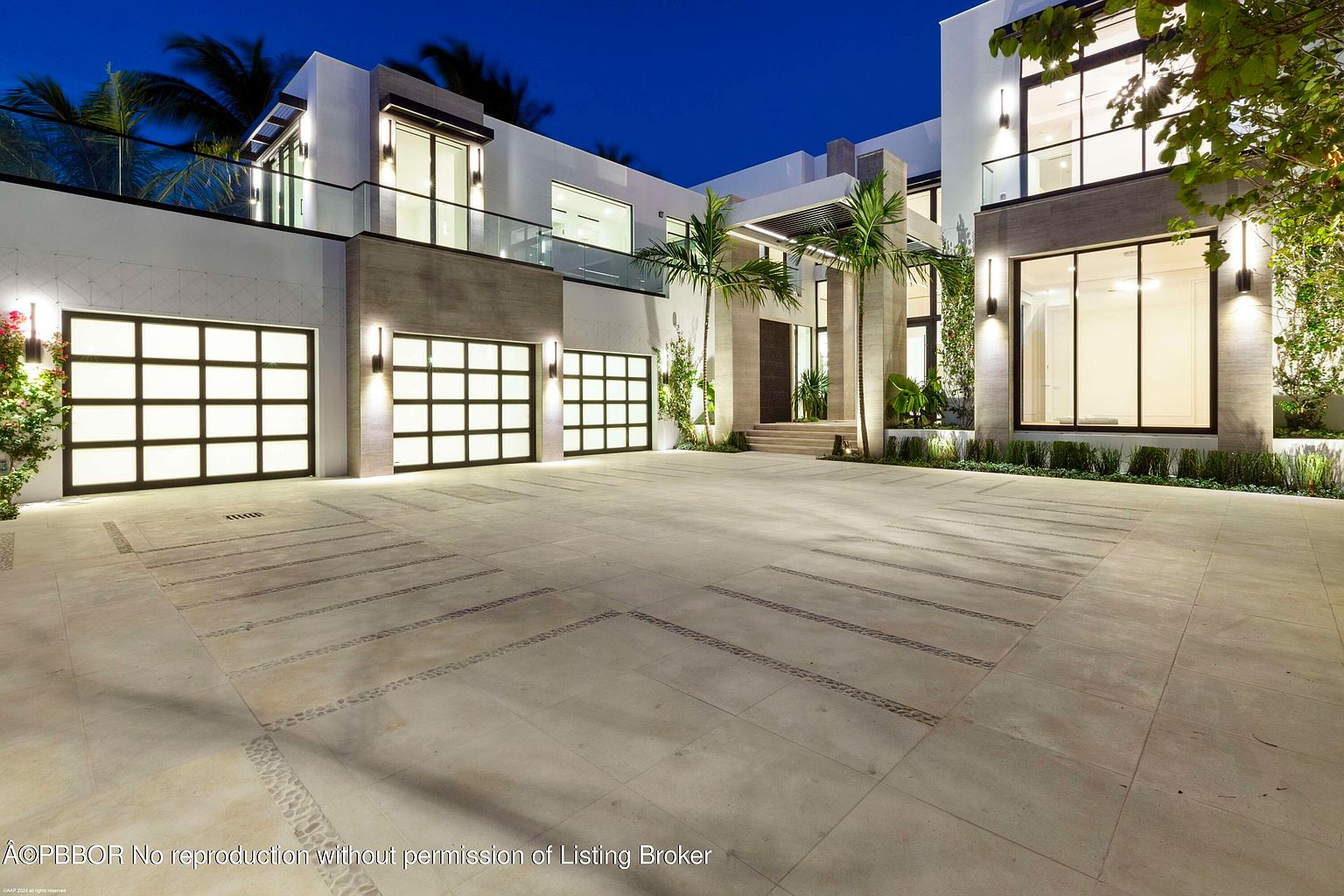 This is a front exterior view of a modern, two-story home at dusk. The house features a three-car garage with frosted glass doors, a stone facade, and large windows. Palm trees and manicured landscaping add to the property's curb appeal, while the driveway is paved with large stone tiles.