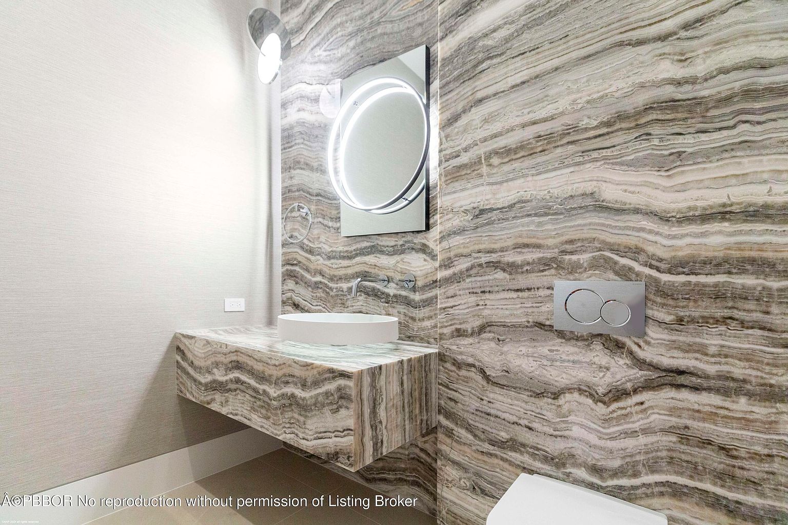 This is a modern bathroom featuring a unique marble wall and countertop. The floating countertop supports a vessel sink, and a circular mirror with integrated lighting adds a contemporary touch. The overall design is sleek and luxurious, creating a spa-like atmosphere.