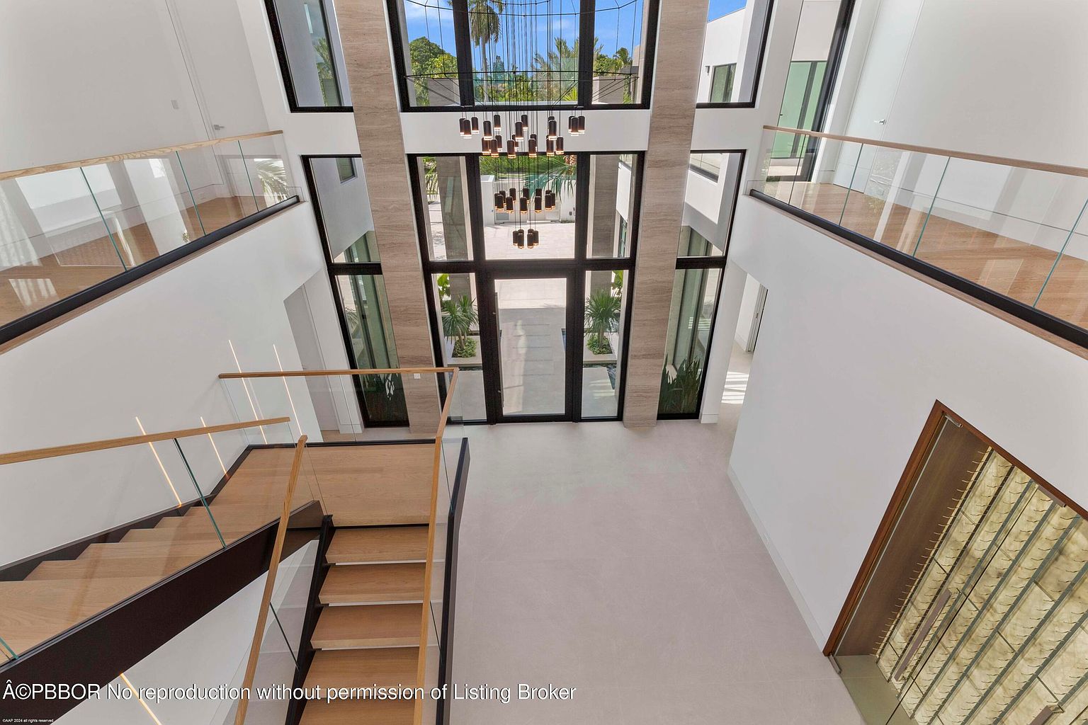 This is an interior shot of a grand foyer featuring a modern staircase with wooden steps and glass railings. The space is illuminated by a contemporary chandelier hanging from the high ceiling, and large windows provide ample natural light and views of the exterior. The overall impression is one of spaciousness and modern elegance.