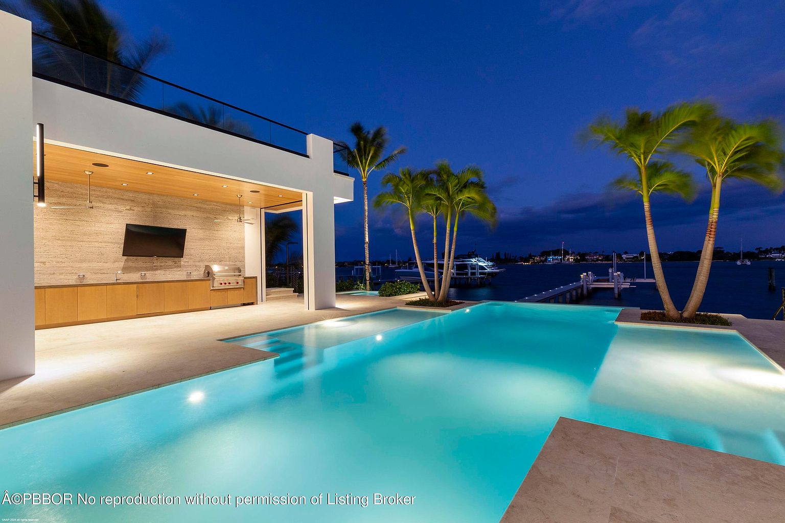 This image showcases a luxurious outdoor pool area at dusk, featuring a modern design with integrated palm trees and a seamless transition to the waterfront. The pool is illuminated, highlighting its turquoise water, and an outdoor kitchen area with a built-in grill and TV is visible in the background. The scene evokes a sense of high-end living and relaxation.
