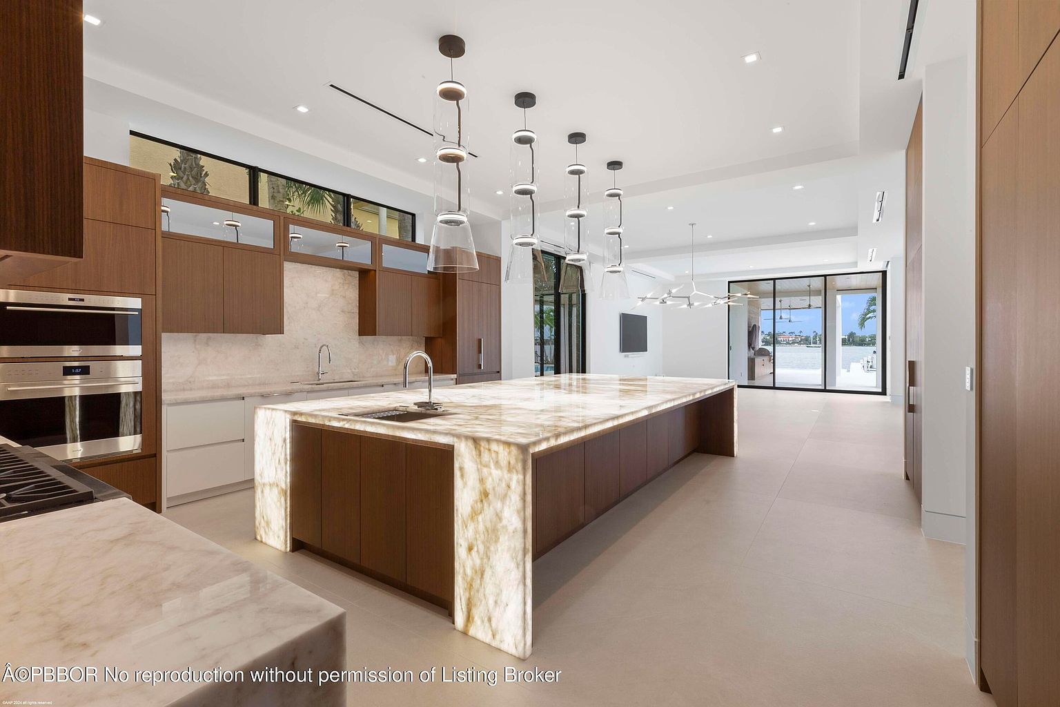 This is a bright and spacious modern kitchen featuring a large island with a marble countertop and waterfall edges, complemented by dark wood cabinetry. The kitchen is well-lit with pendant lights above the island and recessed lighting throughout. Large sliding glass doors offer a view of the waterfront, enhancing the open and airy feel of the space.