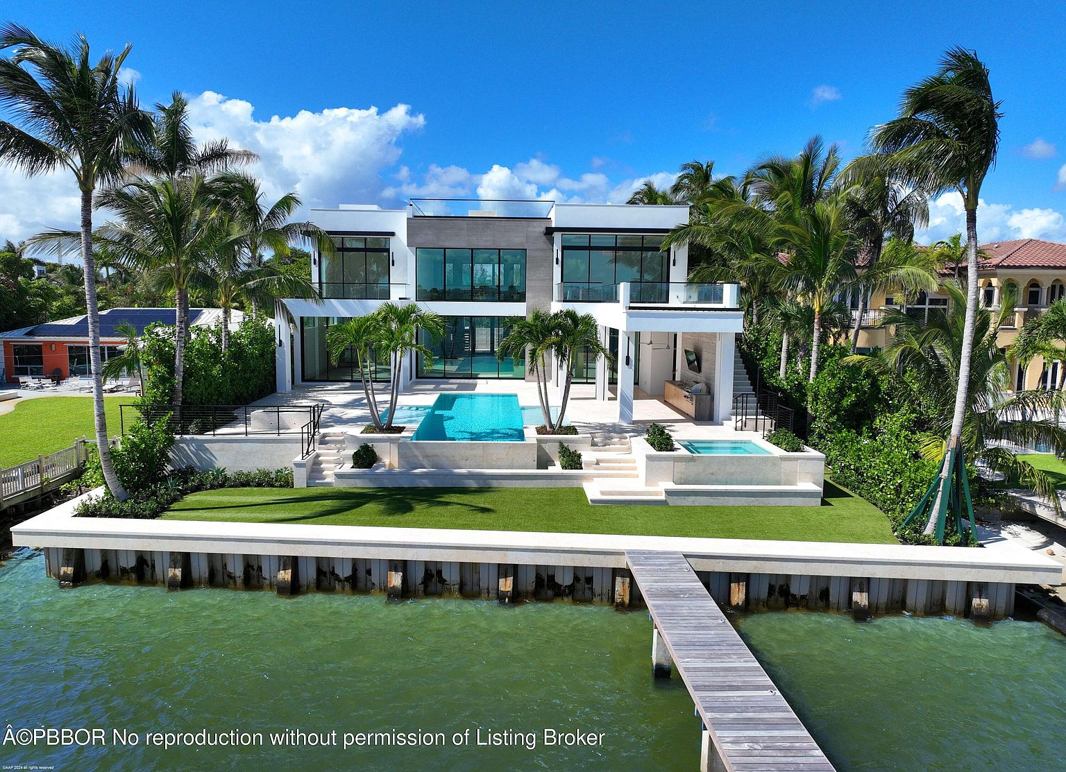 This is a stunning rear view of a modern waterfront home. The property features a sleek, contemporary design with large windows, a swimming pool, a spa, and a private dock. Lush landscaping and palm trees surround the home, creating a tropical oasis.