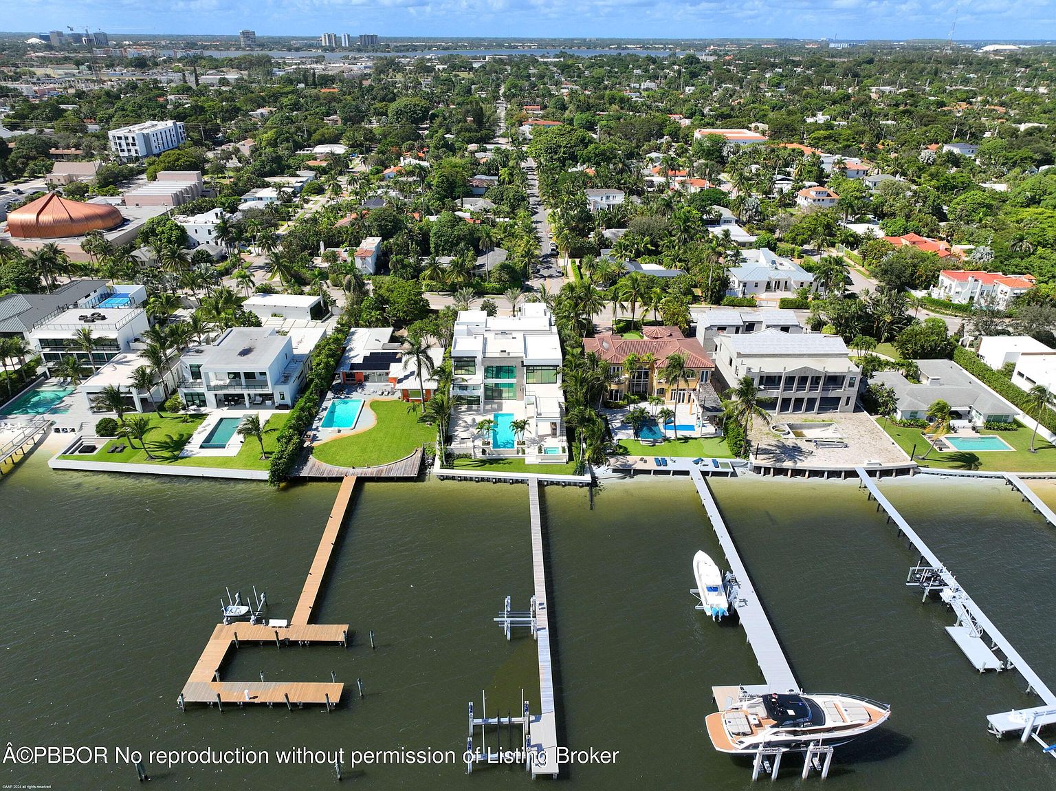 This aerial view showcases waterfront properties with modern architectural designs, featuring private docks and swimming pools. The properties are surrounded by lush greenery and palm trees, offering a luxurious and serene living environment. The perspective is from above, looking down at the properties and the water, providing a comprehensive view of the real estate and its surroundings.
