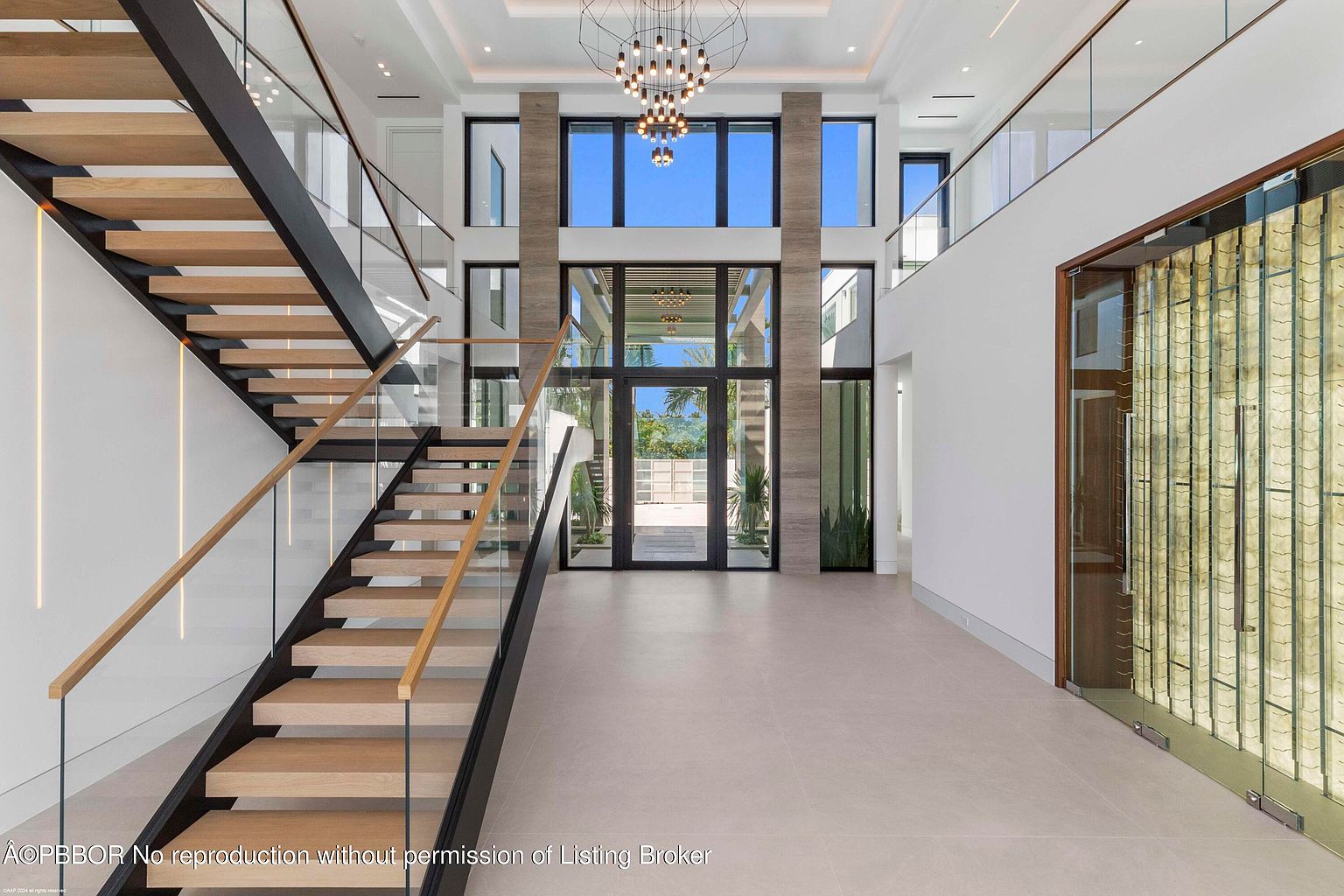 This grand foyer showcases a modern staircase with wooden steps, glass railings, and a dark metal frame. Natural light floods the space through large windows, highlighting the high ceilings and open floor plan. A statement chandelier adds a touch of elegance to the contemporary design.