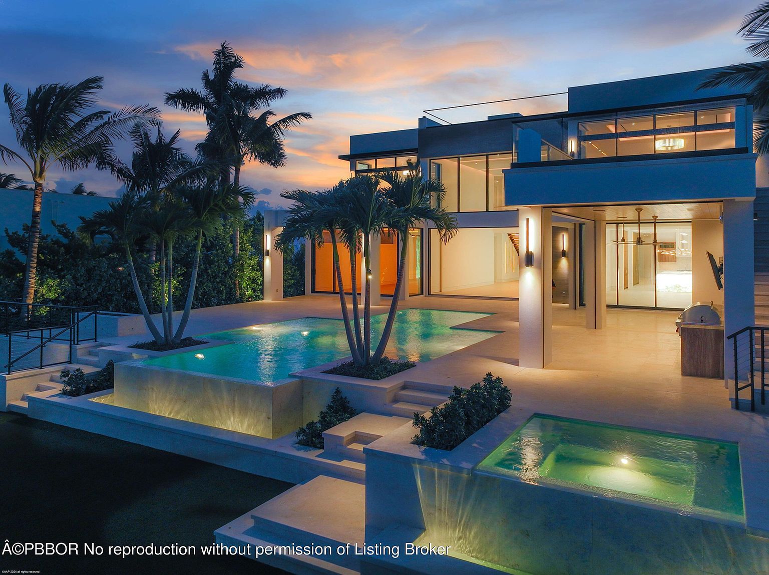 This image showcases the rear exterior of a modern home, emphasizing the luxurious pool and spa area. The multi-tiered pool features integrated planters with palm trees, creating a tropical oasis. The architecture is contemporary with clean lines, large glass windows, and outdoor lighting that enhances the ambiance, perfect for evening relaxation and entertainment.