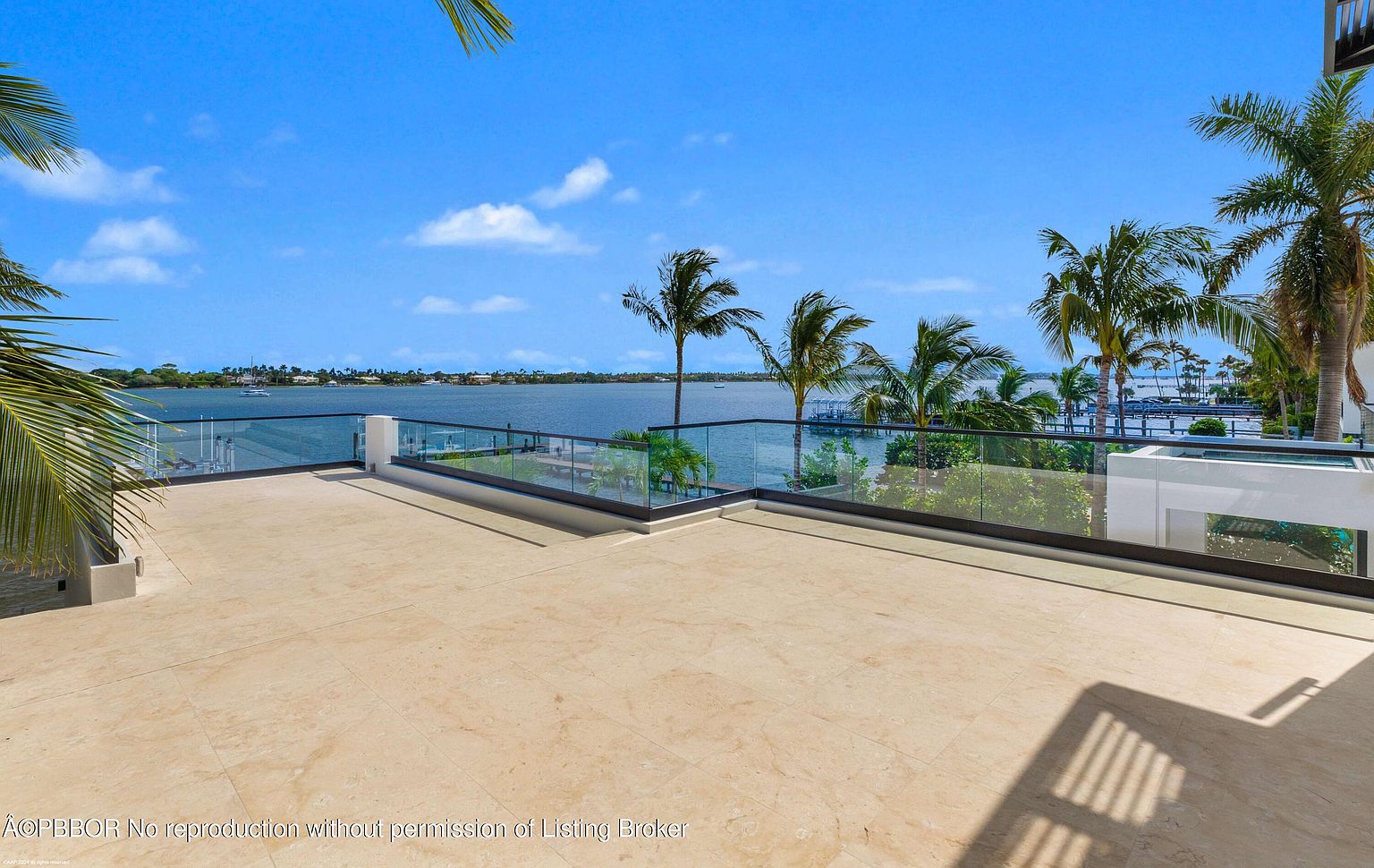 This image showcases a luxurious outdoor patio or deck area with a stunning waterfront view. The space features a light-colored tiled floor, modern glass railings, and lush palm trees in the background, creating a tropical and upscale ambiance. The clear blue sky and calm water enhance the sense of tranquility and exclusivity.