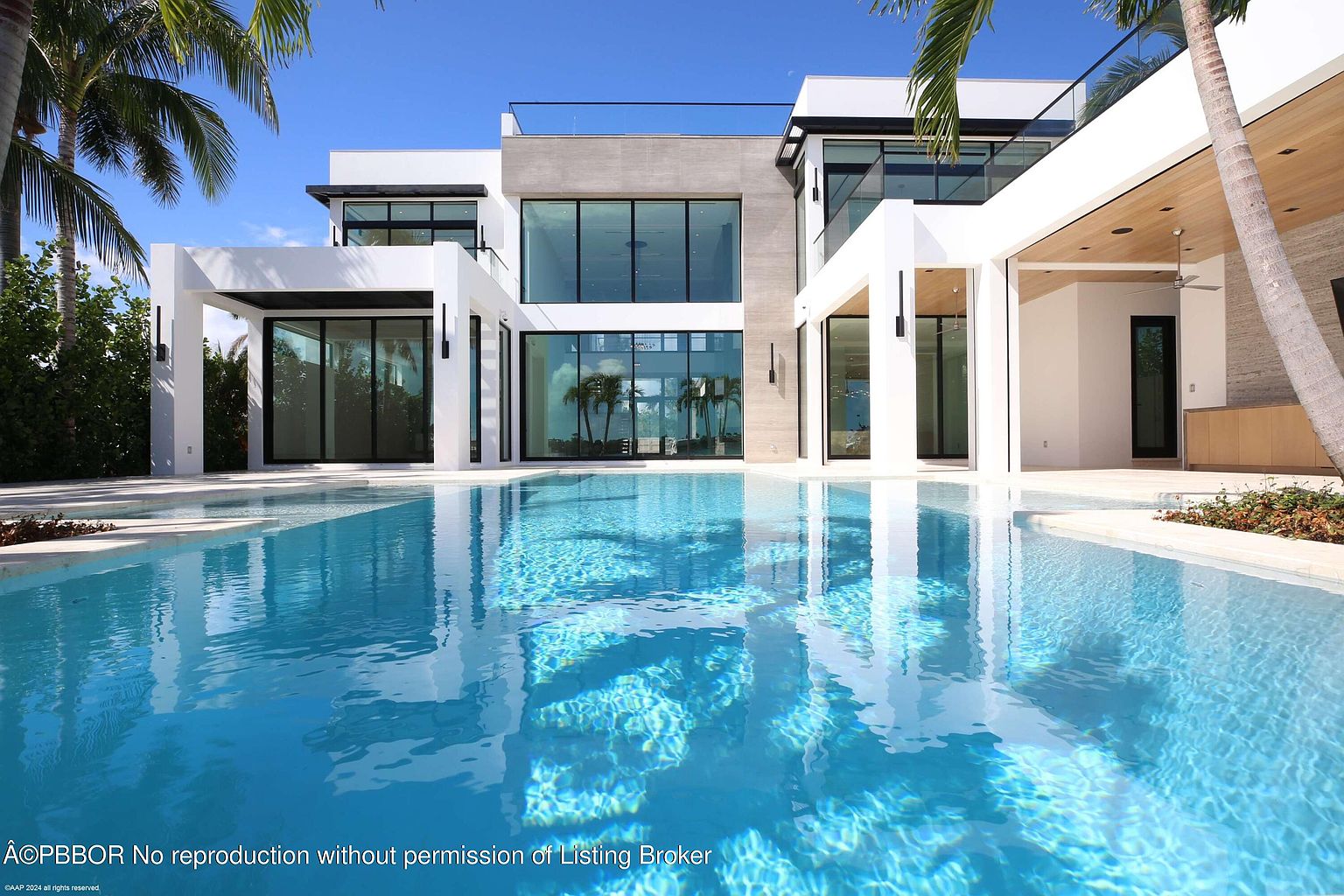 This image showcases the rear exterior of a modern luxury home, emphasizing its stunning pool area. The pool's clear blue water reflects the contemporary architecture of the house, which features large glass windows and white structural elements. Palm trees frame the scene, adding a tropical touch to this inviting outdoor space.