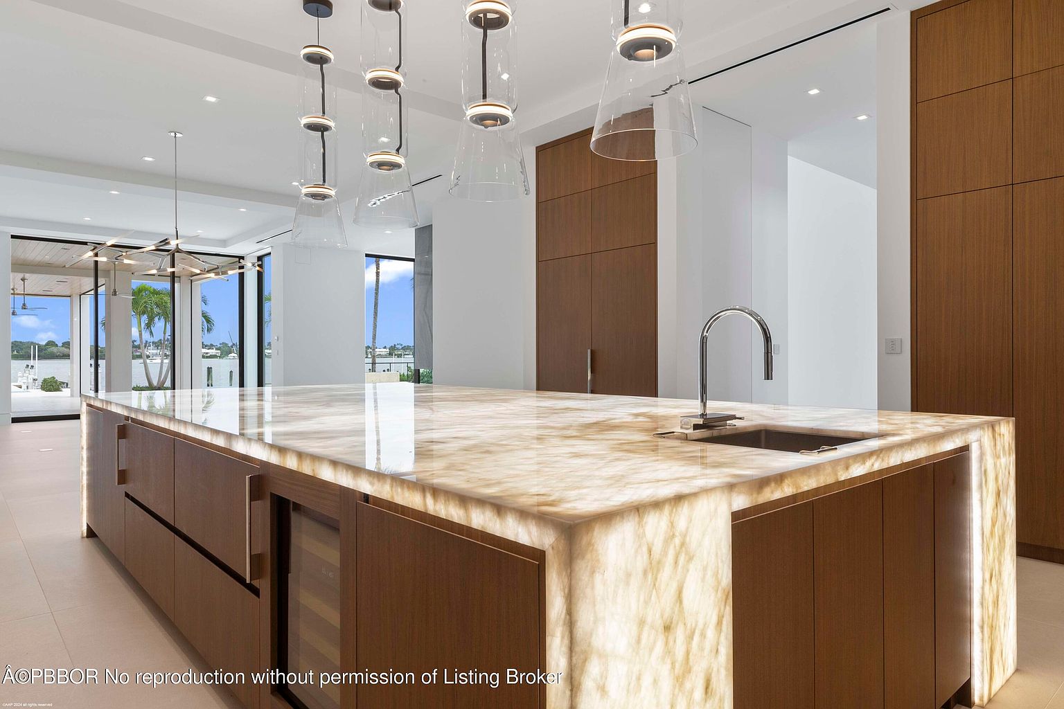 This is a bright and modern kitchen featuring a large island with a unique, illuminated countertop. The cabinetry is a warm wood tone, and pendant lights hang above the island. The kitchen has a clean and luxurious feel, with high-end finishes and a view to the outside.