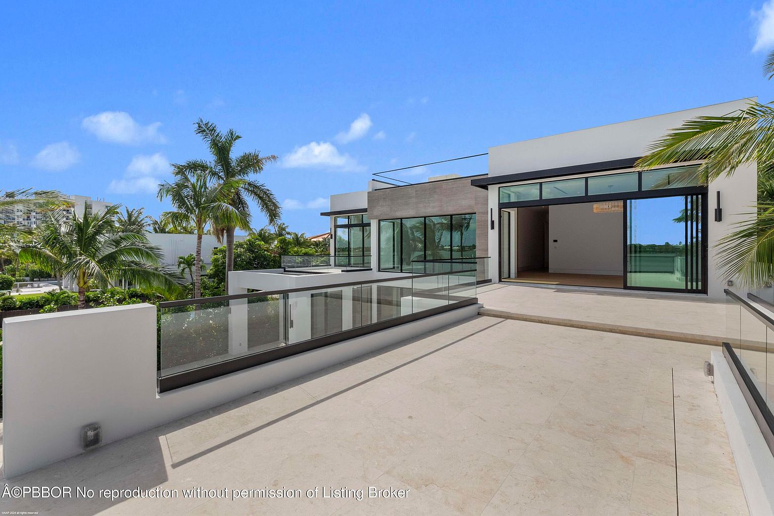 This image showcases a modern home's expansive patio or deck area, featuring sleek glass railings and a light-colored tiled surface. Large sliding glass doors provide seamless indoor-outdoor living, while palm trees and lush greenery add a touch of tropical elegance. The architectural style is contemporary, emphasizing clean lines and open spaces.