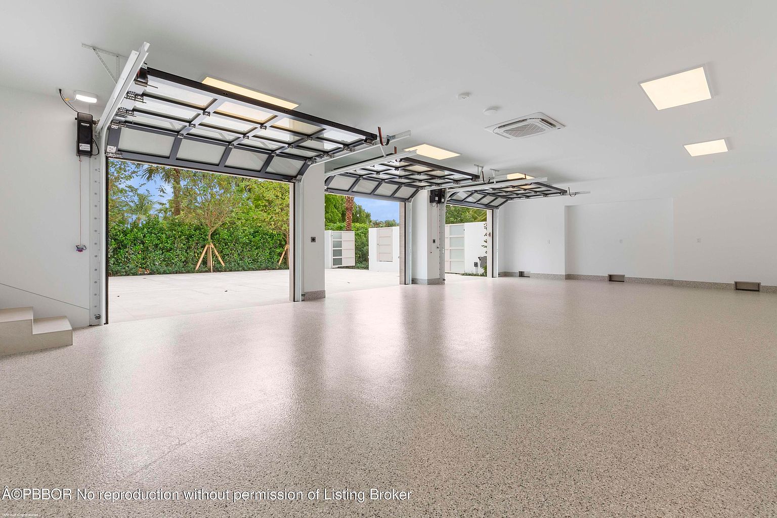 This is a spacious, modern garage with multiple glass-paneled doors that open to the outside. The interior features a light-colored, speckled epoxy floor and white walls, creating a clean and bright space. The open garage doors provide a view of the exterior landscaping, blending indoor and outdoor elements.