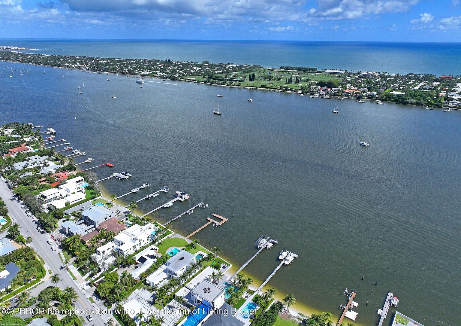This aerial shot showcases a waterfront property with multiple docks extending into the water, accommodating boats. The houses are modern and luxurious, featuring pools and well-manicured lawns. The view extends to a strip of land across the water, dotted with houses and greenery, under a partly cloudy sky.