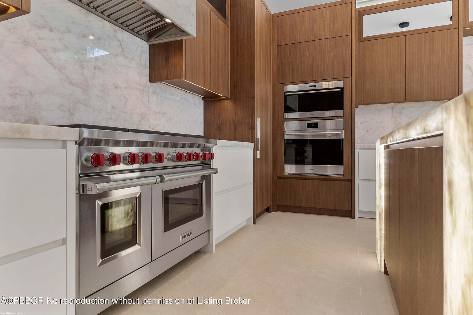 This is a modern kitchen featuring wood cabinets and stainless steel appliances. The focal point is a large Wolf range with red knobs, complemented by built-in ovens. The kitchen has a clean and luxurious feel, with marble backsplash and countertops adding to the high-end aesthetic.
