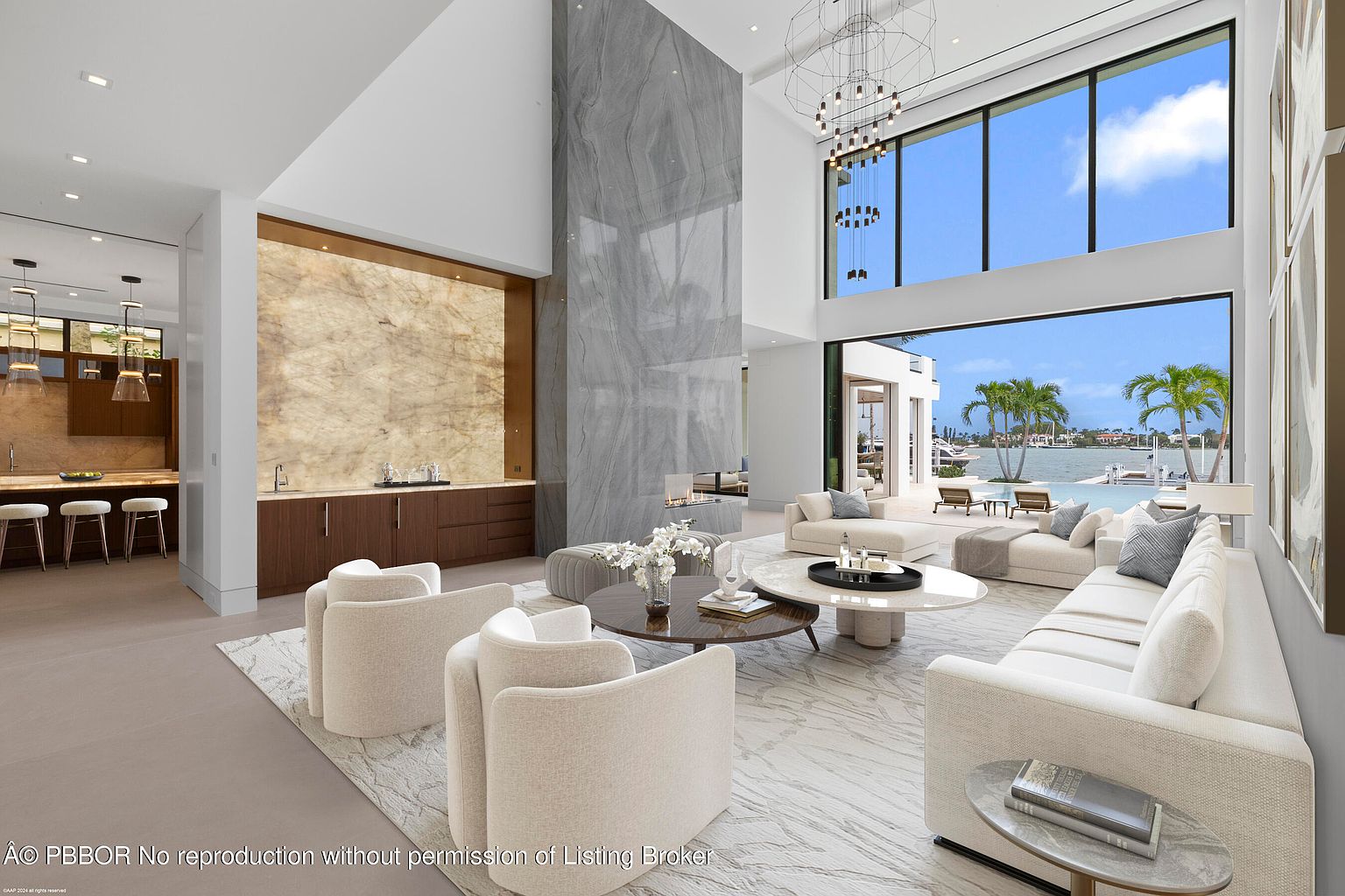 This is a luxurious living room featuring high ceilings, a marble accent wall, and floor-to-ceiling windows offering waterfront views. The room is furnished with modern white sofas, armchairs, and round coffee tables, all arranged on a light-colored patterned rug. The open layout connects to a kitchen area, creating a spacious and inviting atmosphere.
