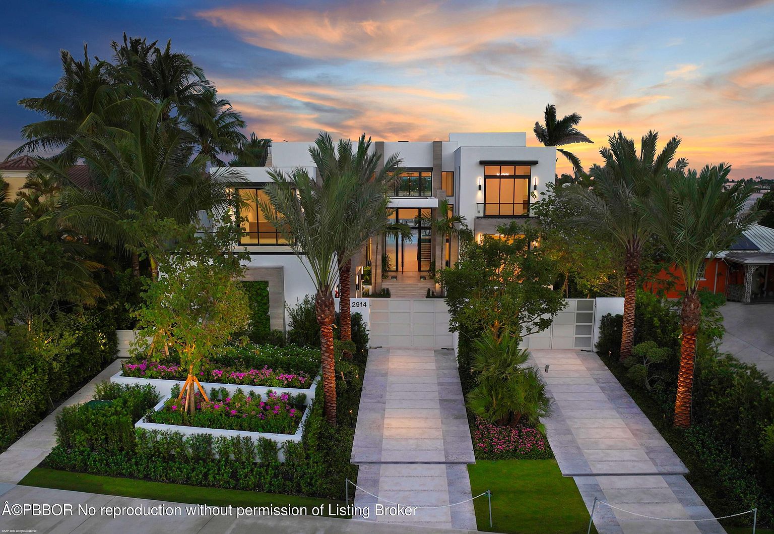 This is a front exterior view of a modern, two-story home with a clean, white facade and large windows. The property features meticulously landscaped gardens with tiered flower beds, palm trees, and a paved driveway leading to a gated entrance. The overall impression is one of luxury and sophisticated design, enhanced by the warm hues of the sunset sky.