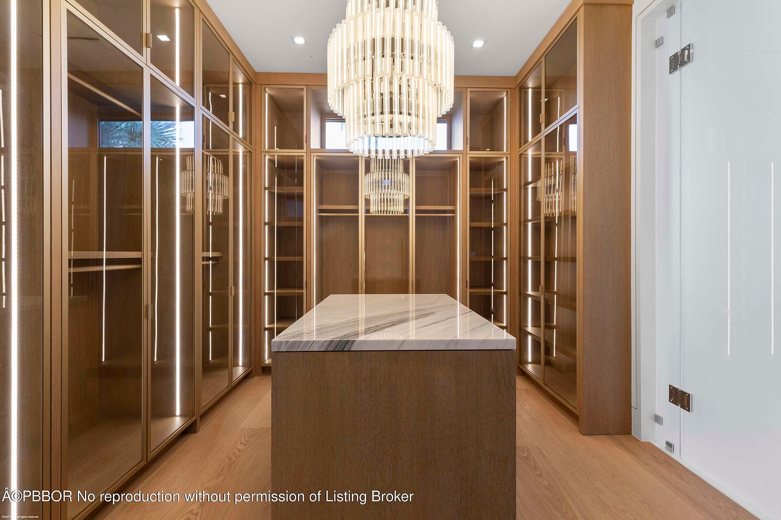 This is a luxurious walk-in closet featuring custom cabinetry with glass doors and integrated lighting. A central island with a marble countertop provides additional storage and display space. The overall design is elegant and sophisticated, creating a high-end dressing area.