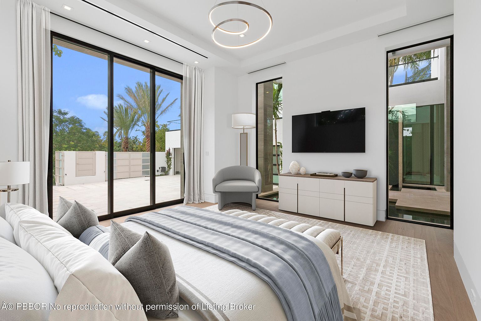This is a bright and airy primary bedroom featuring a large sliding glass door that leads to an outdoor patio. The room is decorated in a modern style with a neutral color palette, a sleek TV console, and a contemporary light fixture. The overall impression is one of luxury and tranquility.