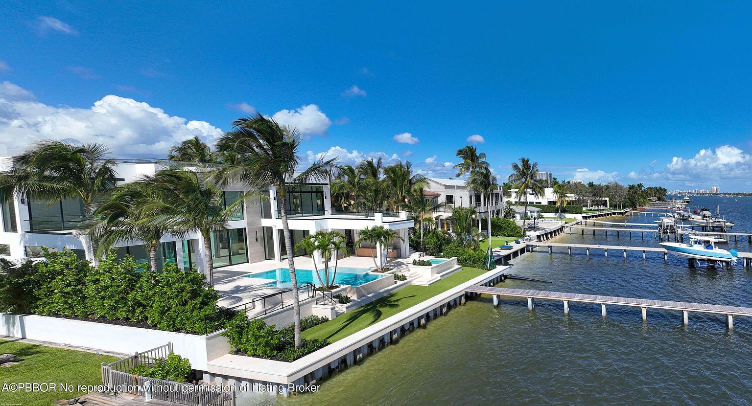 This aerial shot showcases a luxurious waterfront property featuring a modern white house with expansive glass windows and a private pool. Lush palm trees and manicured lawns surround the residence, leading to a private dock with boats. The scene is set against a clear blue sky, emphasizing the property's prime location and upscale appeal.