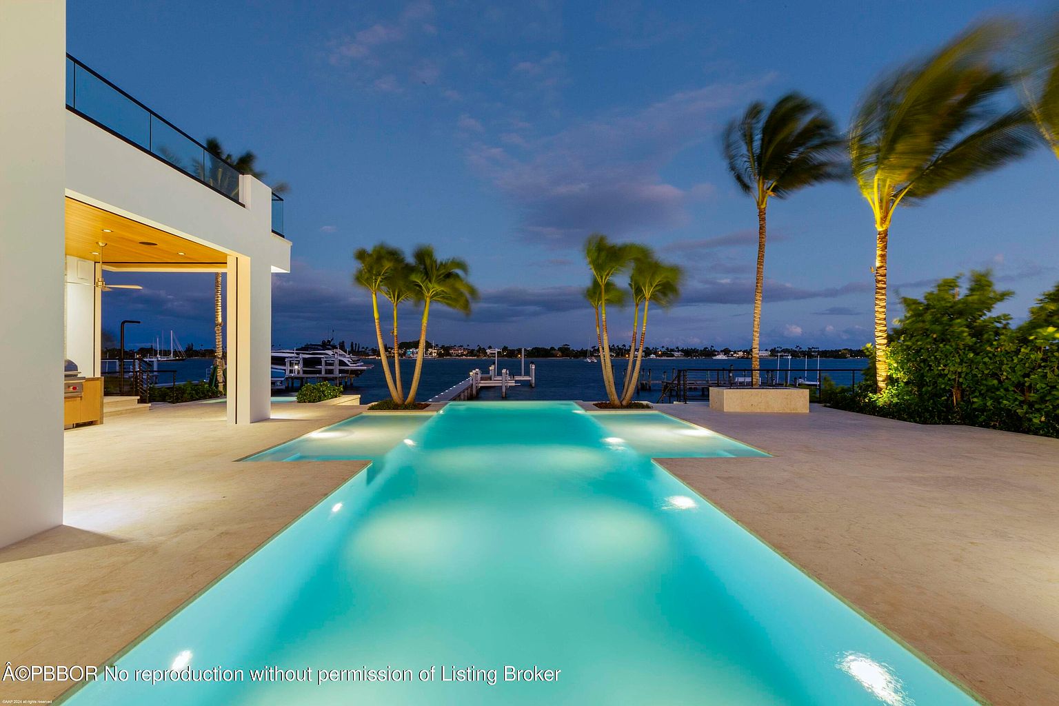 This image showcases a luxurious outdoor pool area at dusk, featuring a modern design with a long, illuminated pool that extends towards a waterfront view. Palm trees line the perimeter, adding a tropical feel, while the architecture of the adjacent building suggests high-end living. The scene evokes a sense of relaxation and upscale leisure.