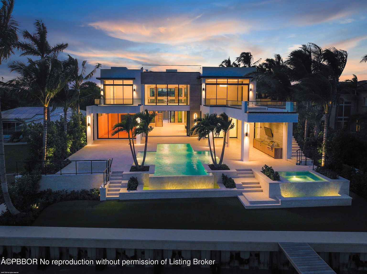This image showcases the rear exterior of a modern luxury home at dusk, featuring a stunning pool and spa area. The architecture emphasizes clean lines and large windows, allowing for ample natural light and views. Palm trees and manicured landscaping enhance the property's appeal, creating a resort-like atmosphere.