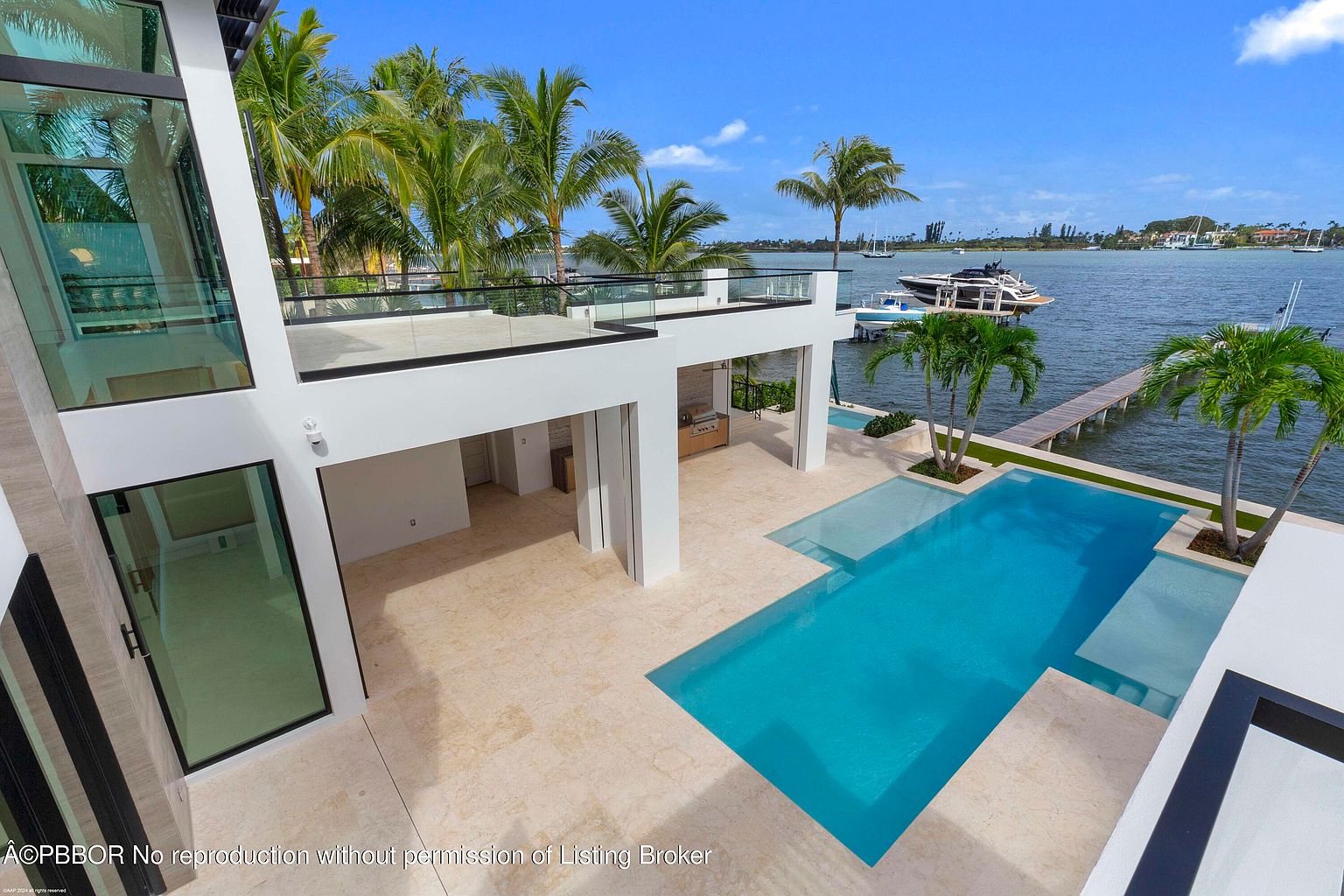 This aerial view showcases a modern luxury home with a stunning waterfront location. The property features a sleek, white exterior, a large swimming pool with integrated steps, and a private dock extending into the water. Palm trees add a tropical touch, and a yacht is moored nearby, emphasizing the upscale lifestyle.