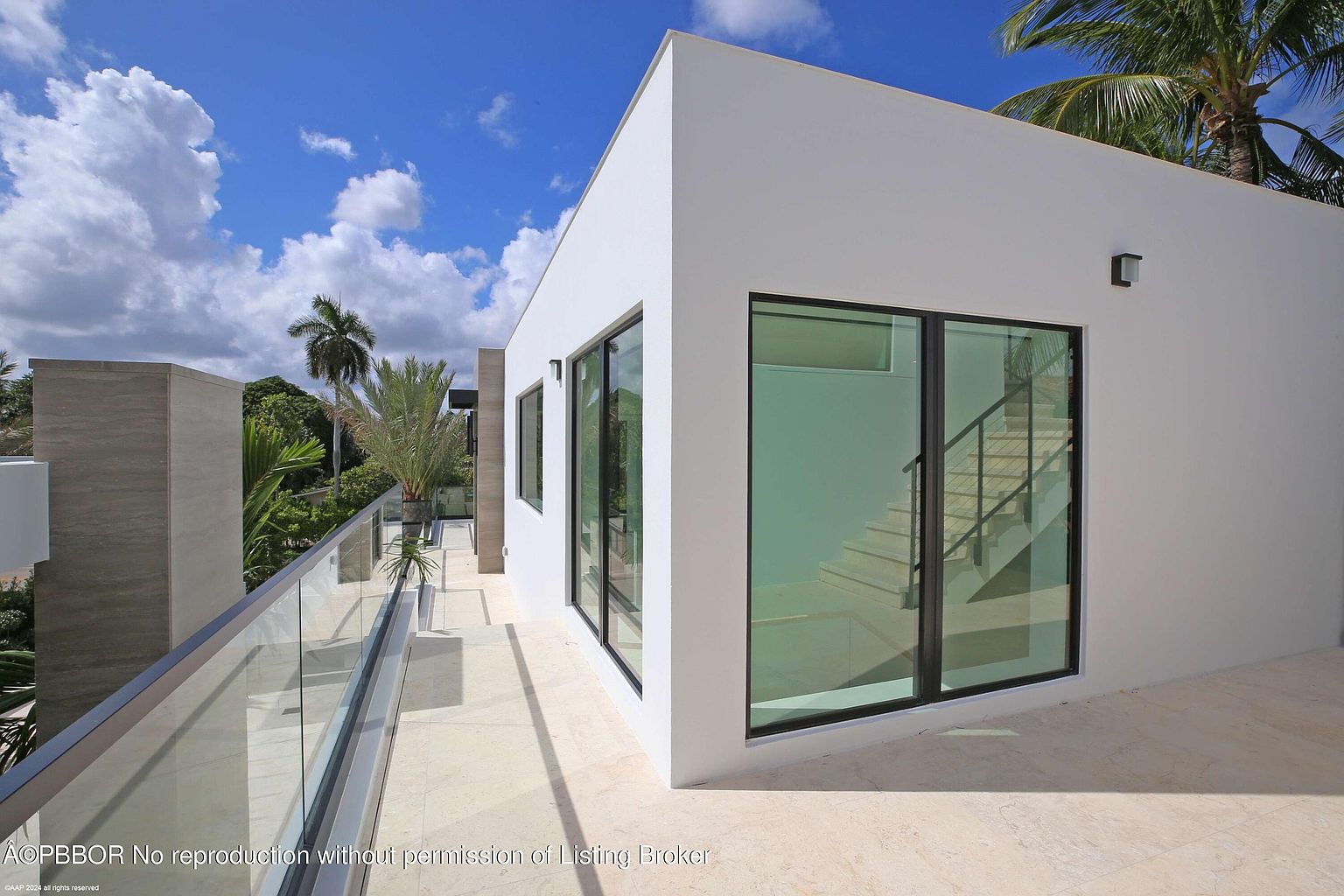 This image showcases a modern balcony or deck area of a contemporary home. The space features a sleek glass railing, light-colored tiled flooring, and large windows offering a glimpse of an interior staircase. The overall design is minimalist and sophisticated, emphasizing clean lines and open space.