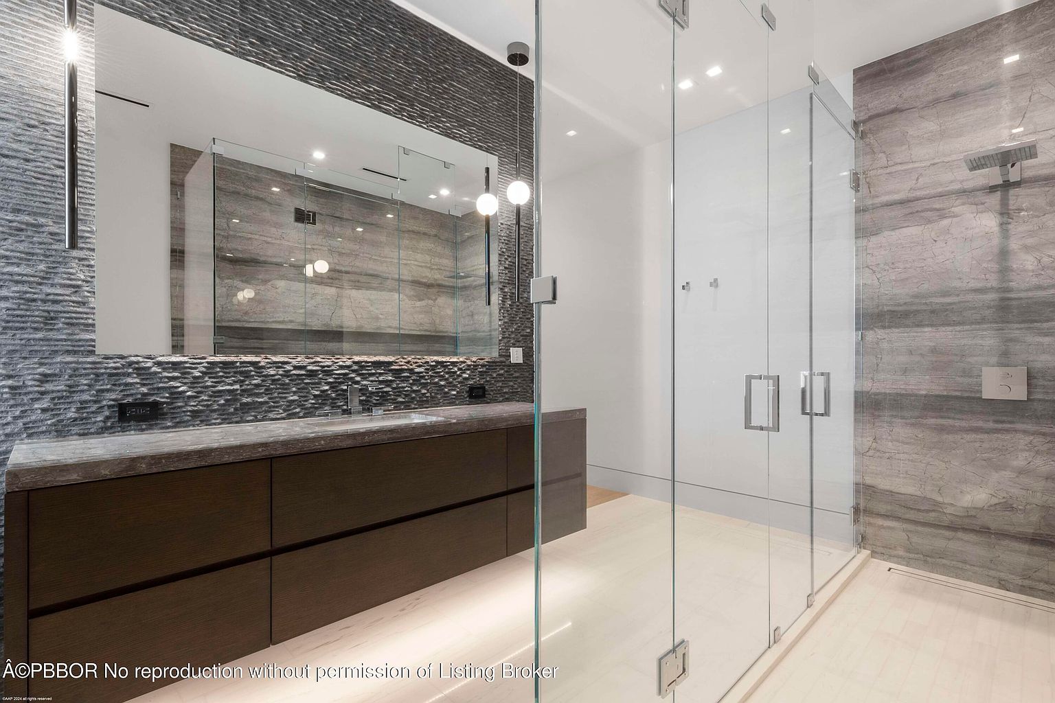 This is a modern primary bathroom featuring a long, dark wood vanity with a gray countertop and a large mirror. The walls are adorned with textured gray tiles, and a glass-enclosed shower with gray marble walls is visible. The overall impression is luxurious and contemporary.