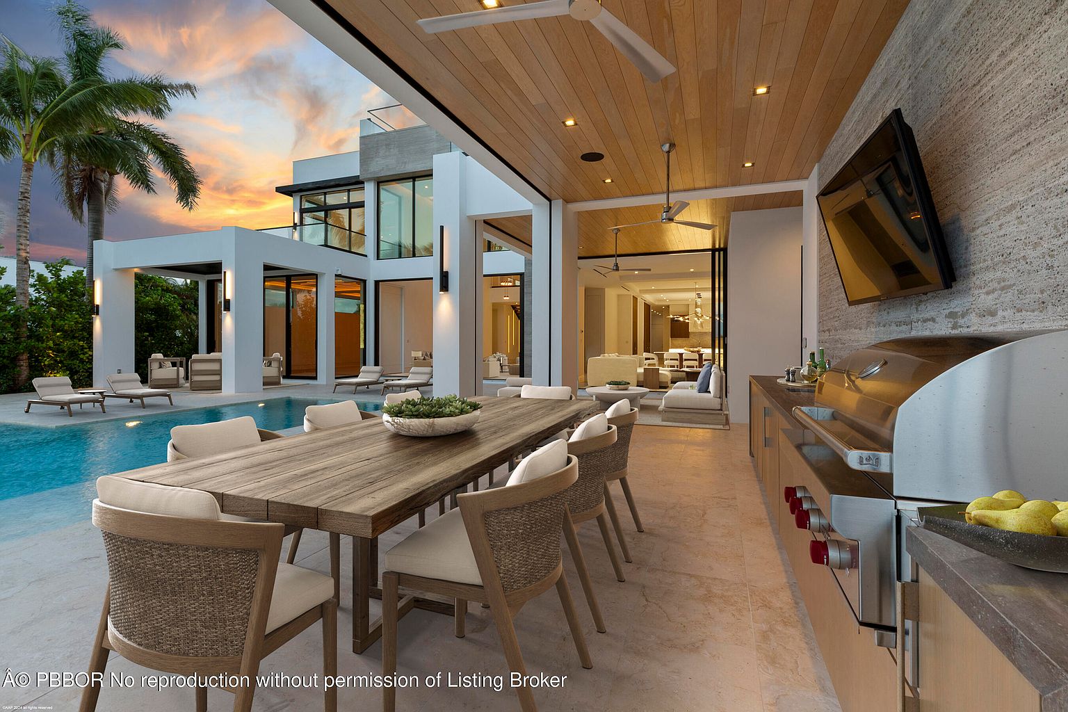 This image showcases a luxurious outdoor patio area with a dining table set for eight, a built-in grill, and a large flat-screen TV. The patio is adjacent to a modern home with floor-to-ceiling windows and a swimming pool, creating a seamless indoor-outdoor living experience. The warm lighting and sunset sky enhance the inviting and upscale ambiance.