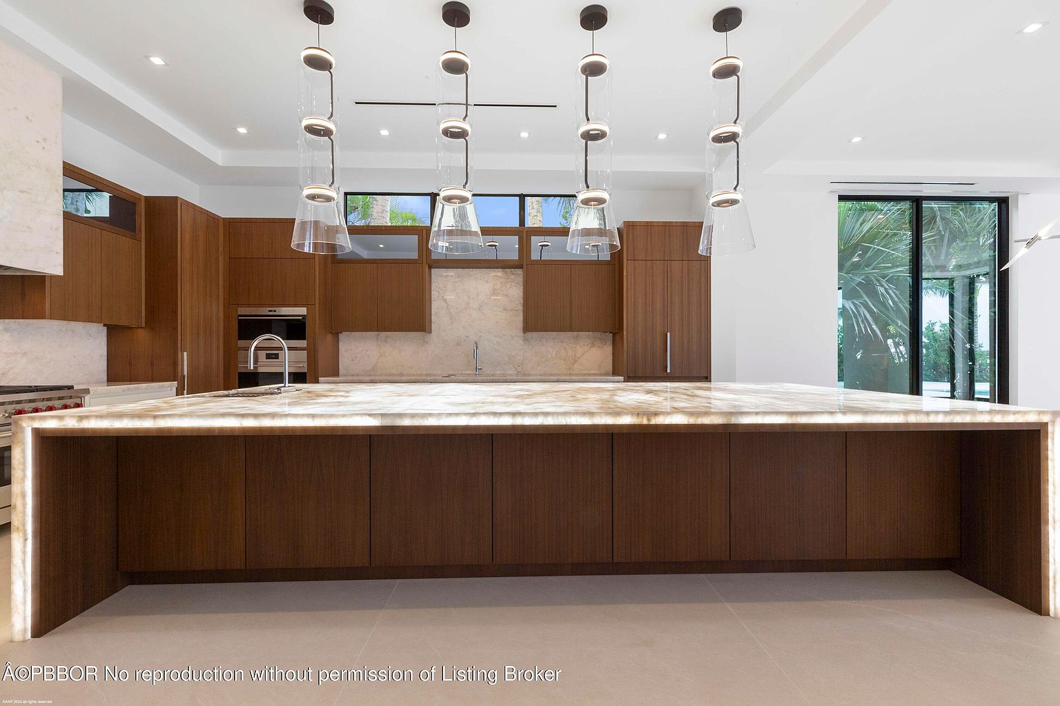 This is a modern kitchen featuring sleek, wood-paneled cabinetry and a large, illuminated island with a marble countertop. The kitchen is well-lit with unique pendant lighting fixtures hanging above the island. A window provides natural light and a glimpse of the outdoors, enhancing the contemporary and luxurious feel of the space.