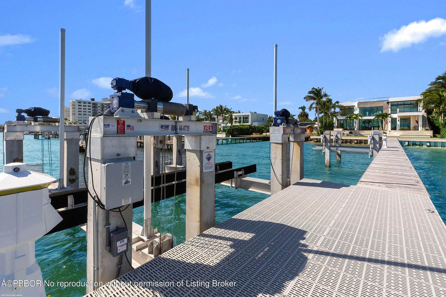 This image showcases a modern waterfront property with a private dock and boat lift. The dock features a gray, textured surface and leads to a contemporary home with large windows and clean lines. The scene is set against a clear blue sky, highlighting the property's access to the water and its modern architectural design.