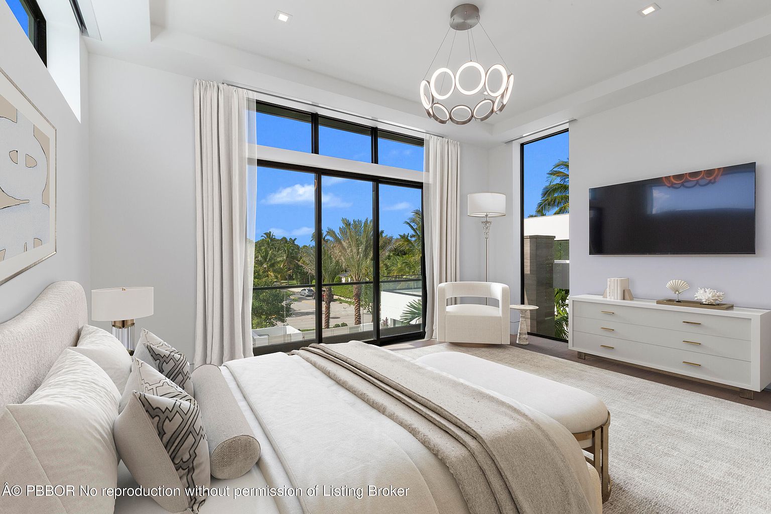 This is a bright and airy primary bedroom featuring a large bed with white linens and decorative pillows. A wall of windows offers a view of lush greenery, while a modern chandelier hangs from the ceiling. A white dresser with a television above it adds a touch of luxury.