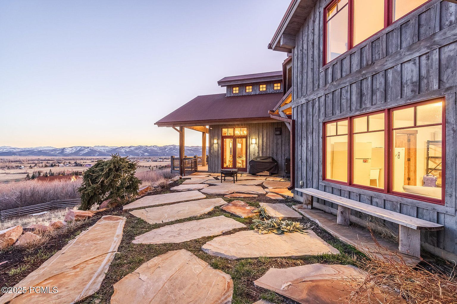 This image showcases the rear exterior of a rustic-style home, featuring a stone pathway leading to a covered patio area with outdoor seating and a grill. The house is clad in weathered wood siding with red-framed windows, and a bench sits against the house. The landscape includes natural stone elements and a view of distant mountains, creating a serene and inviting atmosphere.
