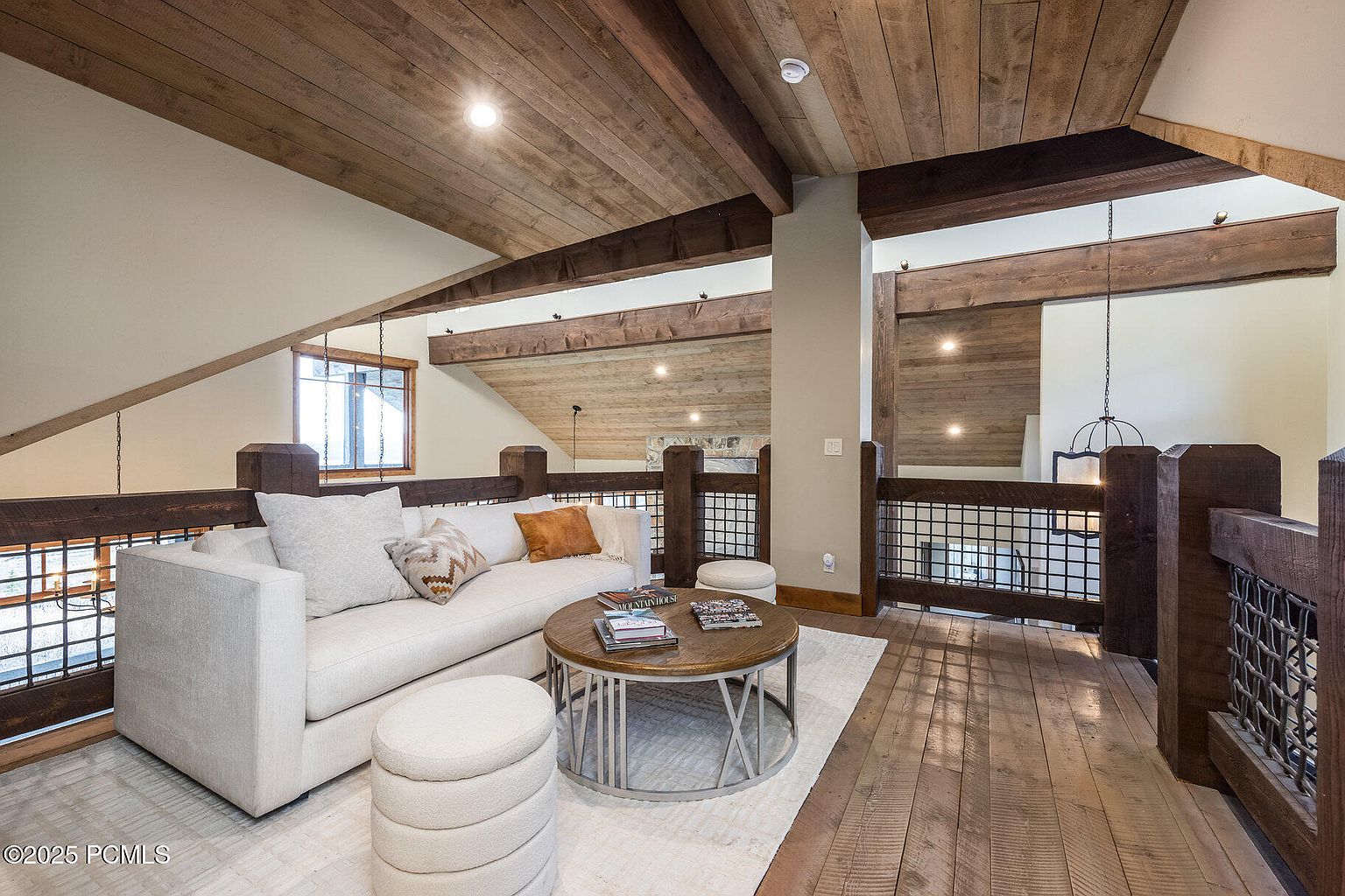 This interior shot showcases a cozy living room area on an upper level, featuring a comfortable white sofa with decorative pillows, a round wooden coffee table, and two matching ottomans. The room is defined by its rustic wooden ceiling and railing, complemented by a light-colored rug and hardwood flooring, creating a warm and inviting atmosphere.