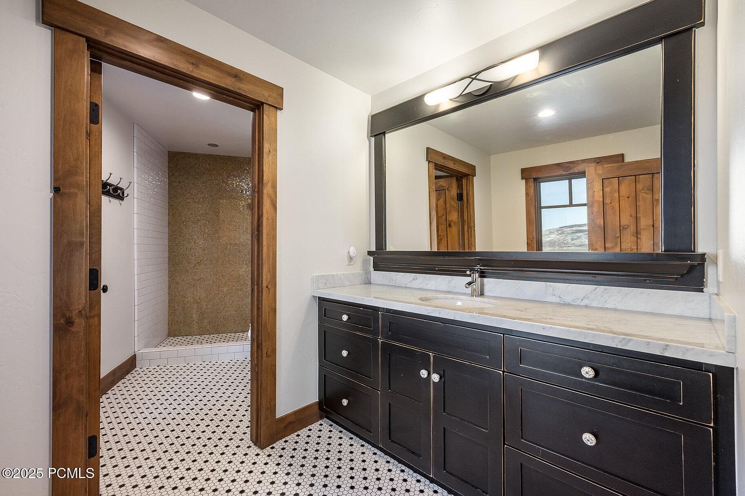 This is a well-appointed primary bathroom featuring a dark wood vanity with a marble countertop and a large framed mirror. The floor is tiled with a classic black and white pattern, and a wooden-framed doorway leads to a shower with mosaic tile accents. The overall impression is one of rustic elegance and functionality.