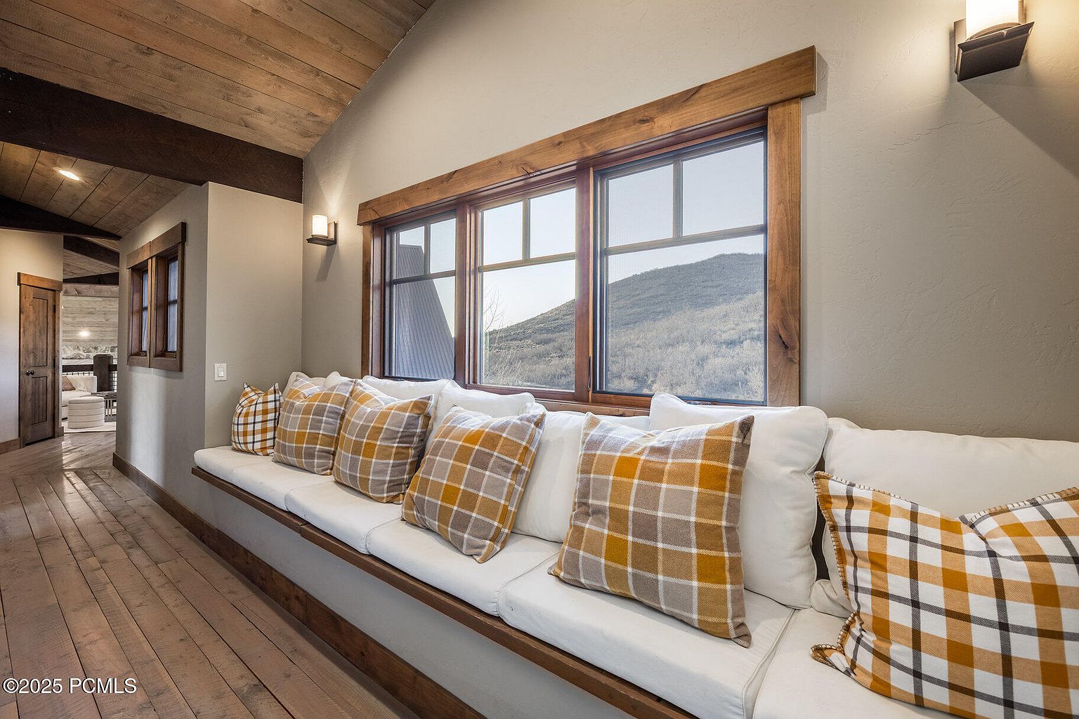This interior shot showcases a hallway with a built-in window seat adorned with white cushions and plaid pillows, creating a cozy and inviting space. The hallway features wooden floors and a wood-paneled ceiling, adding a rustic touch to the home's design. Natural light streams through the windows, highlighting the architectural details and the warm color palette.