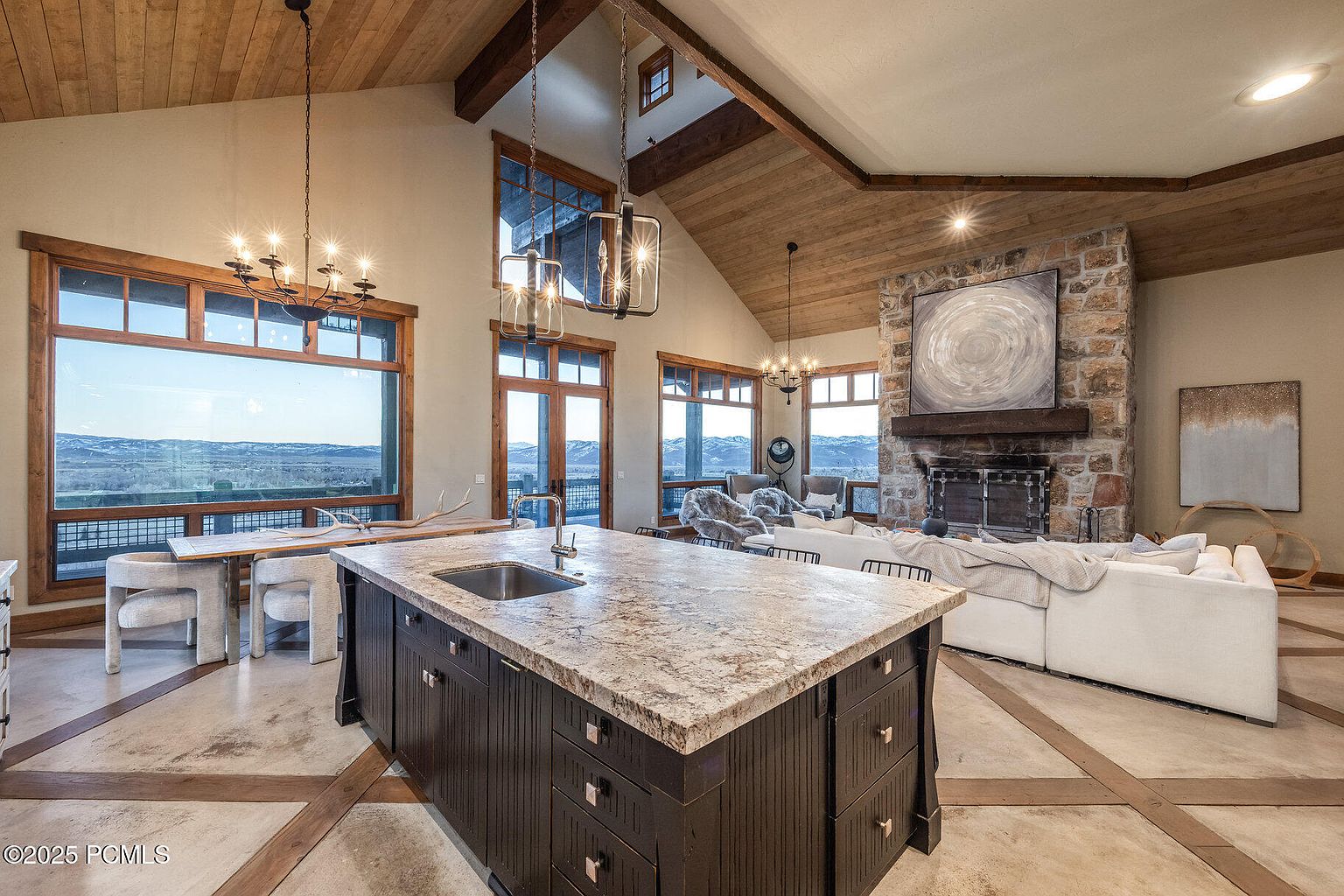 This interior shot showcases a luxurious living room and kitchen area with a rustic yet elegant design. The kitchen features a large island with a stone countertop and dark wood cabinetry, while the living room boasts a stone fireplace and comfortable white sectional sofa. Large windows offer stunning views of the landscape, and the wood-paneled ceiling adds warmth and character to the space.