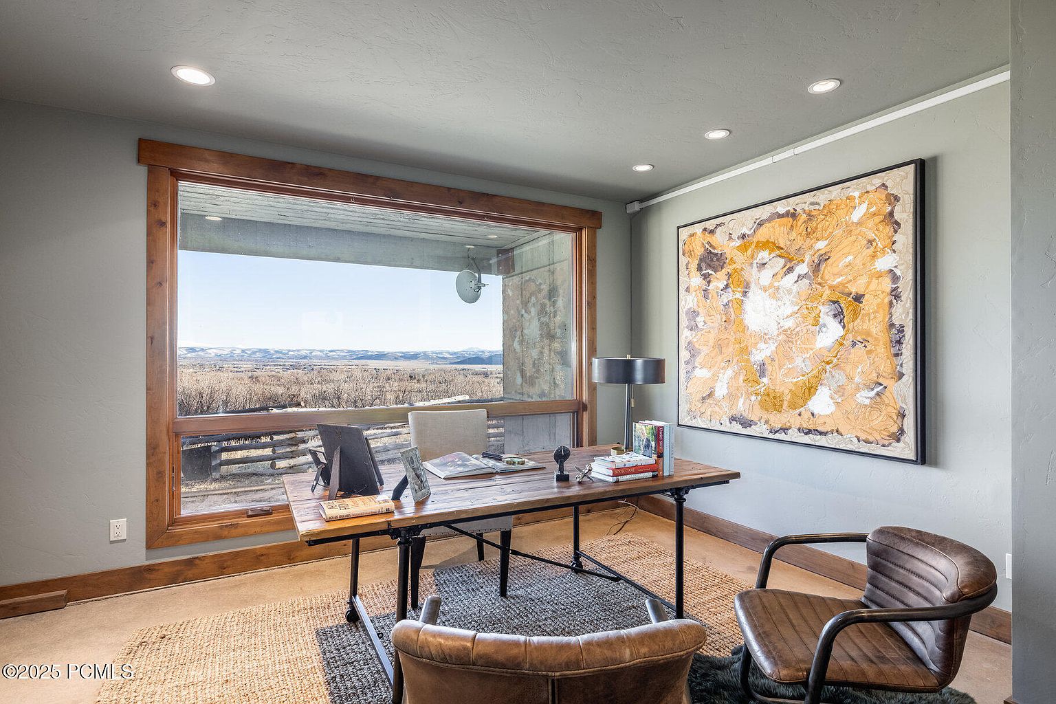 This is an interior shot of a home office featuring a rustic wooden desk with black metal legs, positioned in front of a large window offering a scenic view of a landscape. The room is decorated with a large abstract painting and a leather chair, complemented by a jute rug and recessed lighting, creating a warm and inviting workspace.