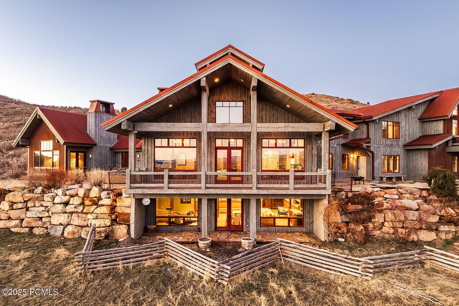 This is a front view of a large, rustic-style home with a red roof and wooden exterior. The house features a prominent balcony with red-framed windows and doors, and a stone foundation that blends into the landscape. The property is surrounded by a natural, grassy yard with a wooden fence, creating a sense of privacy and connection to nature.