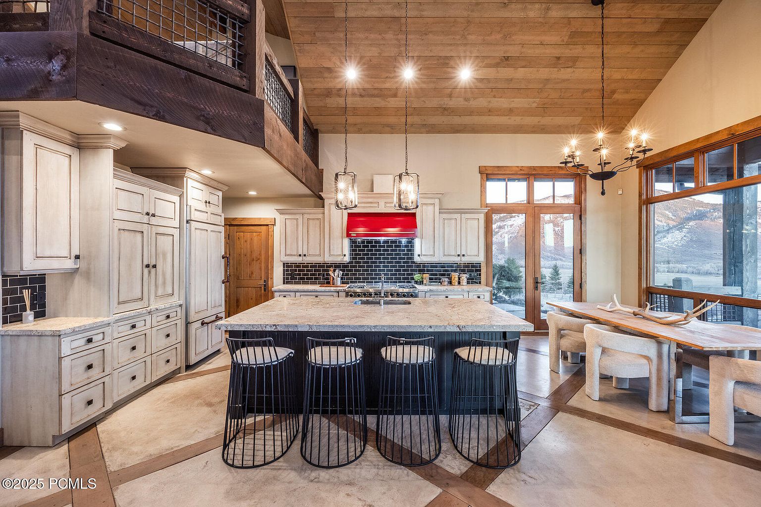 This is a well-lit kitchen featuring a large island with seating, custom cabinetry, and a black subway tile backsplash. The kitchen also has a red range hood and stainless steel appliances. The open floor plan connects the kitchen to the dining area, which has a large window with a view of the mountains.