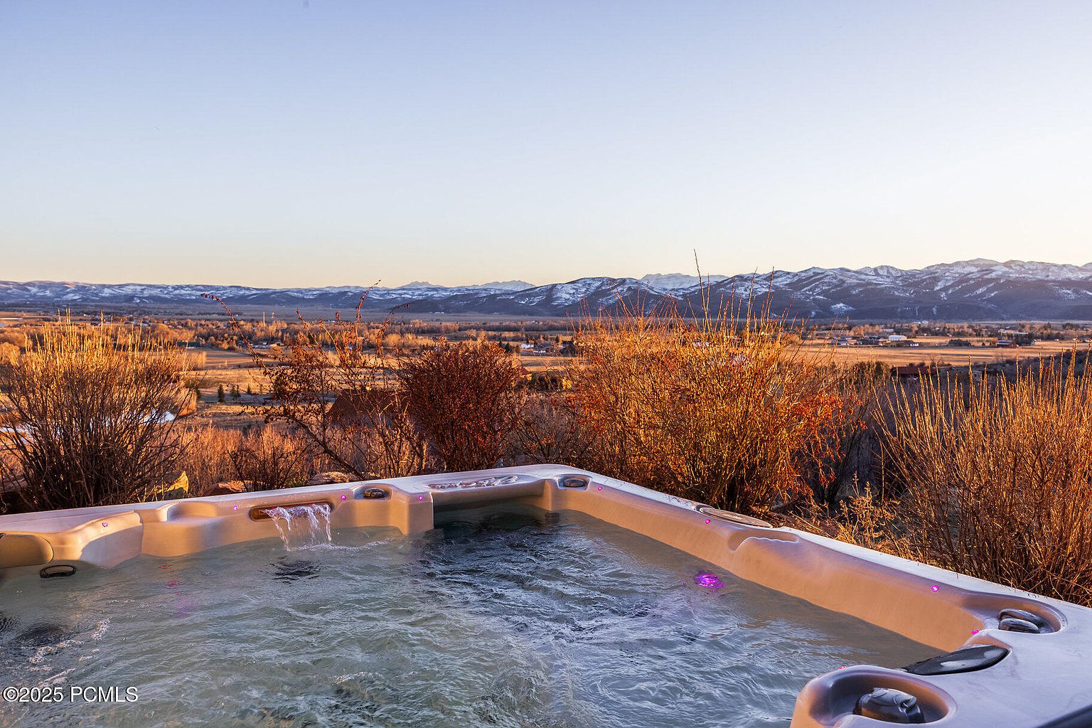 The image showcases a luxurious hot tub with a scenic mountain view. The hot tub features built-in lighting and a waterfall feature, creating a relaxing ambiance. The surrounding landscape includes golden fields and distant snow-capped mountains, enhancing the property's appeal for relaxation and outdoor enjoyment.