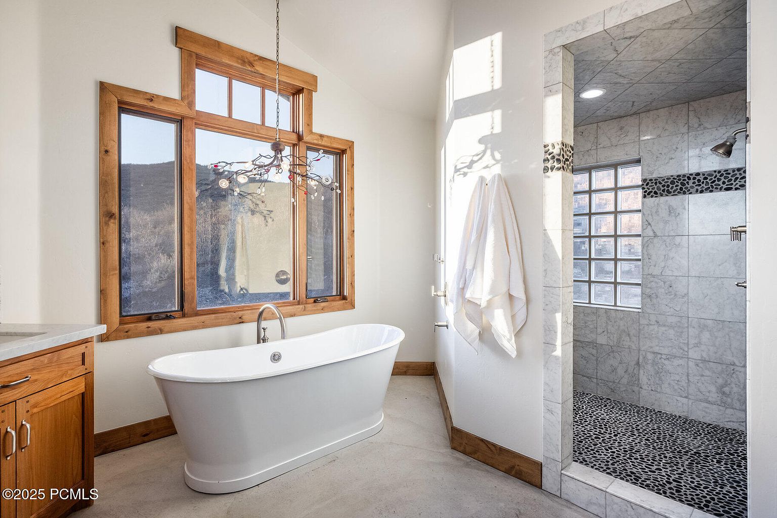 This is a primary bathroom featuring a freestanding white bathtub positioned beneath a large window with wood trim. The shower is enclosed with marble and glass blocks, showcasing a pebble floor. The room has a modern yet rustic aesthetic with a neutral color palette and natural light.