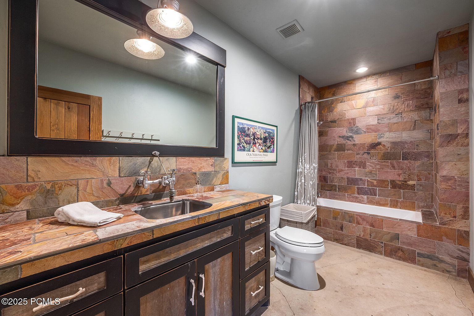 This is a bathroom featuring a stone-tiled shower and wall behind the sink. The vanity has a dark wood frame with rustic wood doors and drawers, topped with a stone countertop and a stainless steel sink. A large mirror hangs above the sink, and a toilet sits next to the shower, with a framed picture on the wall above it.