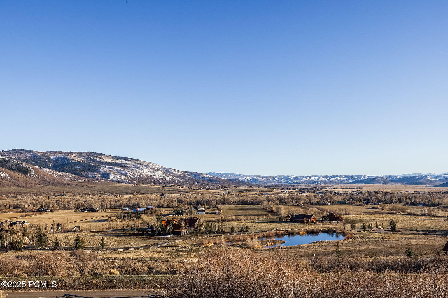This aerial view showcases a sprawling landscape with scattered homes and a serene pond in the foreground. The scene is framed by rolling hills and distant mountains under a clear blue sky, creating a sense of tranquility and vastness. The overall impression is one of rural charm and peaceful living.