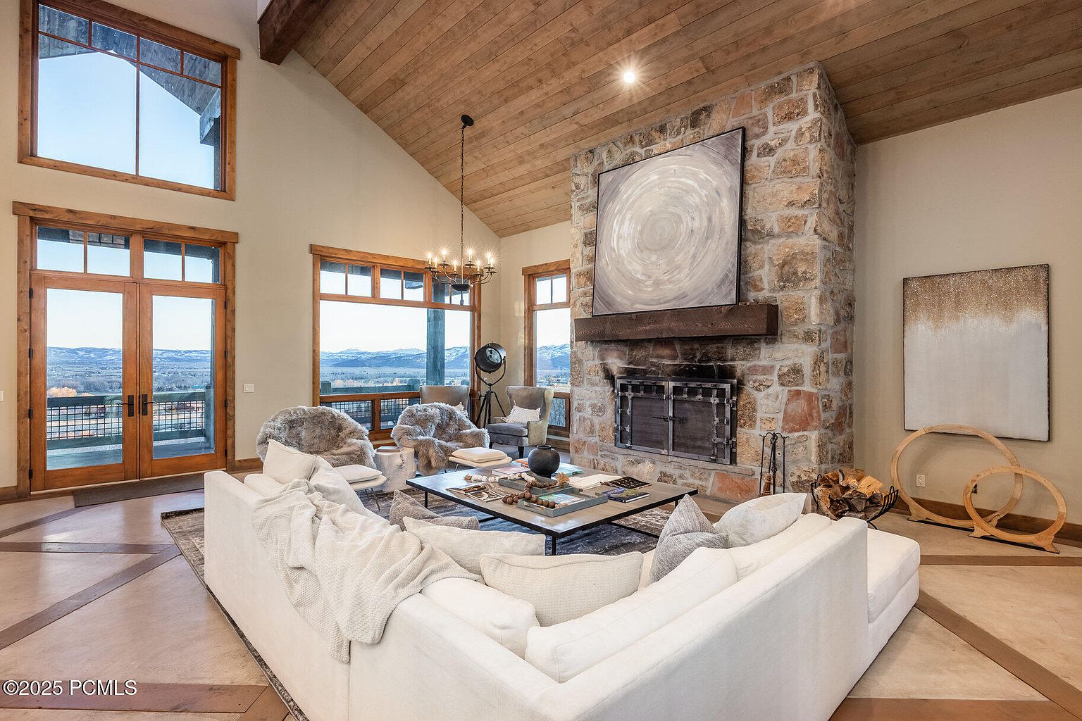 This is an interior shot of a luxurious living room featuring a stone fireplace as a focal point, complemented by a large abstract painting above the mantel. The room is furnished with a comfortable white sectional sofa, cozy armchairs, and a stylish coffee table, all bathed in natural light from the large windows offering scenic views. The wooden ceiling and flooring add warmth and character to the space, creating an inviting and sophisticated atmosphere.